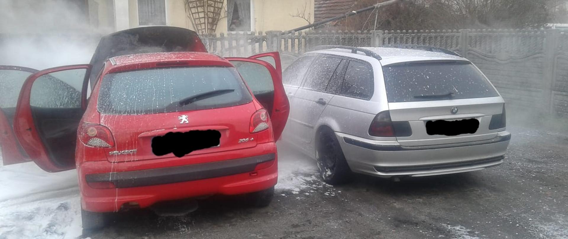 Przed posesją stoją dwa samochody osobowe. Od strony lewej zdjęcia jedne z nich to czerwony Peugeot z otwartymi czterema drzwiami, a obok w odległości jednego metra stoi srebrny samochód osobowy typu kombi marki BMW. Z czerwonego samochodu marki Peugeot z przedniej części to jest komory silnika wydobywa się para, która powstała w wyniku gaszenia pianą gaśniczą przez Państwową straż pożarną. Z lewej strony czerwonego samochodu na ziemi znajduje się rozlewisko z piany gaśniczej