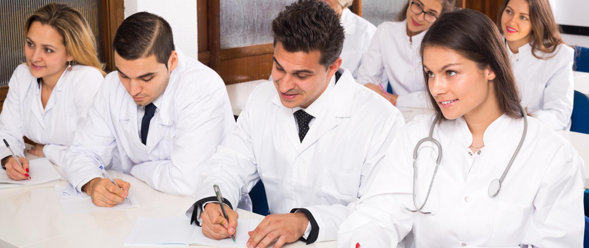 Zdjęcie ilustracyjne, przedstawiające młodych lekarzy robiących notatki przy biurkach.