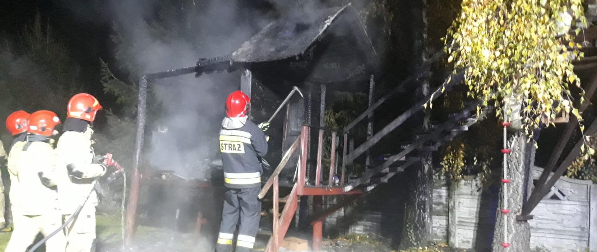 Zdjęcie przedstawia czterech strażaków w ubraniach specjalnych z czerwonymi hełmami na głowach, którzy przystępują do rozbiórki nadpalonych elementów drewnianego domku dla dzieci. Zdjęcie zrobione w porze nocnej.