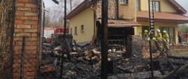 Na pierwszym planie widać za ogrodzeniem posesji zwęglone drewno, zgliszcza po budynku drewnianym dobudowany do murowanego zbudowanego z czerwonej cegły. Na drugim planie uwidoczniony jest budynek mieszkalny z żółtą elewacją i czerwoną dachówką. Można zauważyć że część elewacji oraz dachu uległo spaleniu. Strażacy będący przed budynkiem sprawiają drabinę aluminiową. W tle widać czerwony wóz strażacki, zachmurzone niebo oraz drzewo.