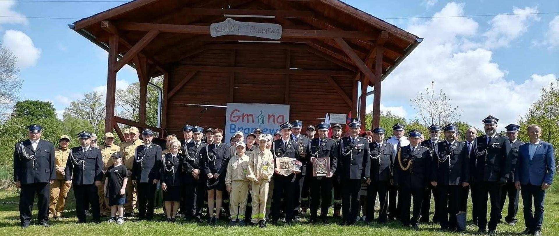 Na tle drewnianej wiaty stoją strażacy w mundurach galowych. Słonecznie.