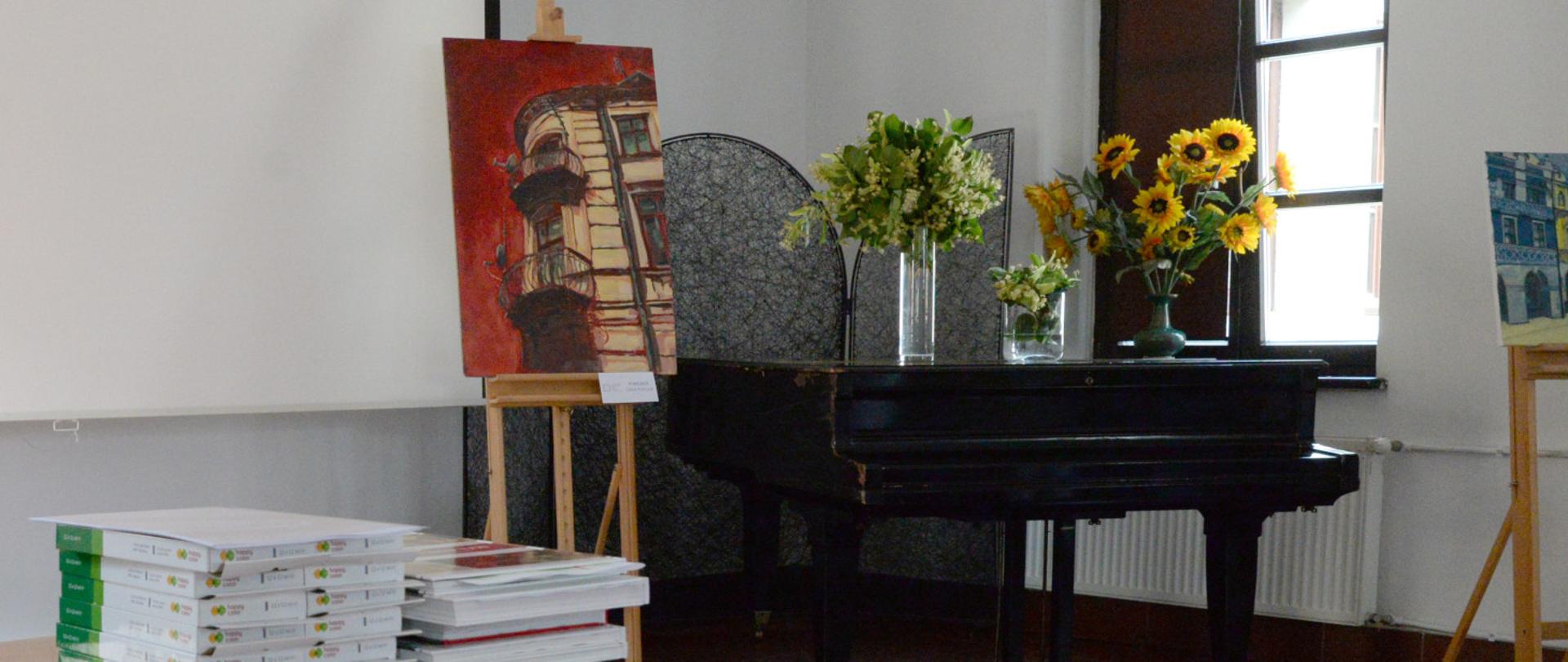 po lewej książki na stoliku, centralnie fortepian a na nim wazony z kwiatami, obok sztaluga z obrazem