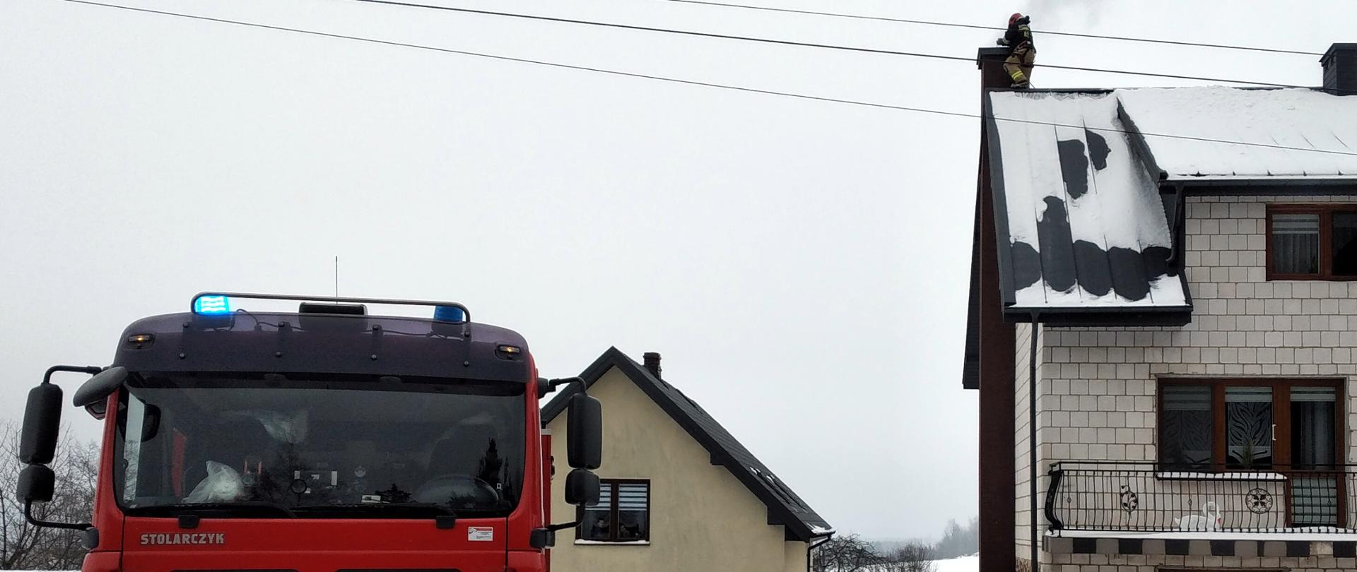 Zdjęcie przedstawia budynek jednorodzinny w miejscowości Malmurzyn 28 z samochodem pożarniczym GCBA 5/32 z JRG nr 2 w Kielcach na pierwszym planie. W oddali przy kominie znajduje się strażak. Z komina wydobywa się lekki dym