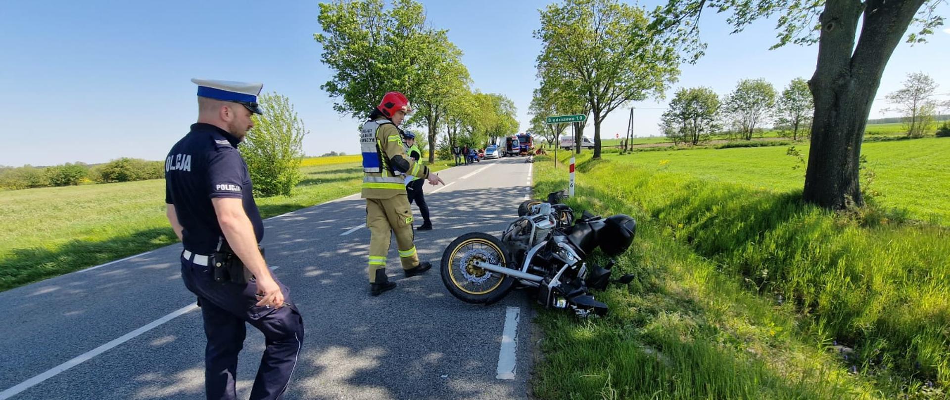 Widać uszkodzony motocykl na poboczu drogi, w pobliżu strażak i policjant