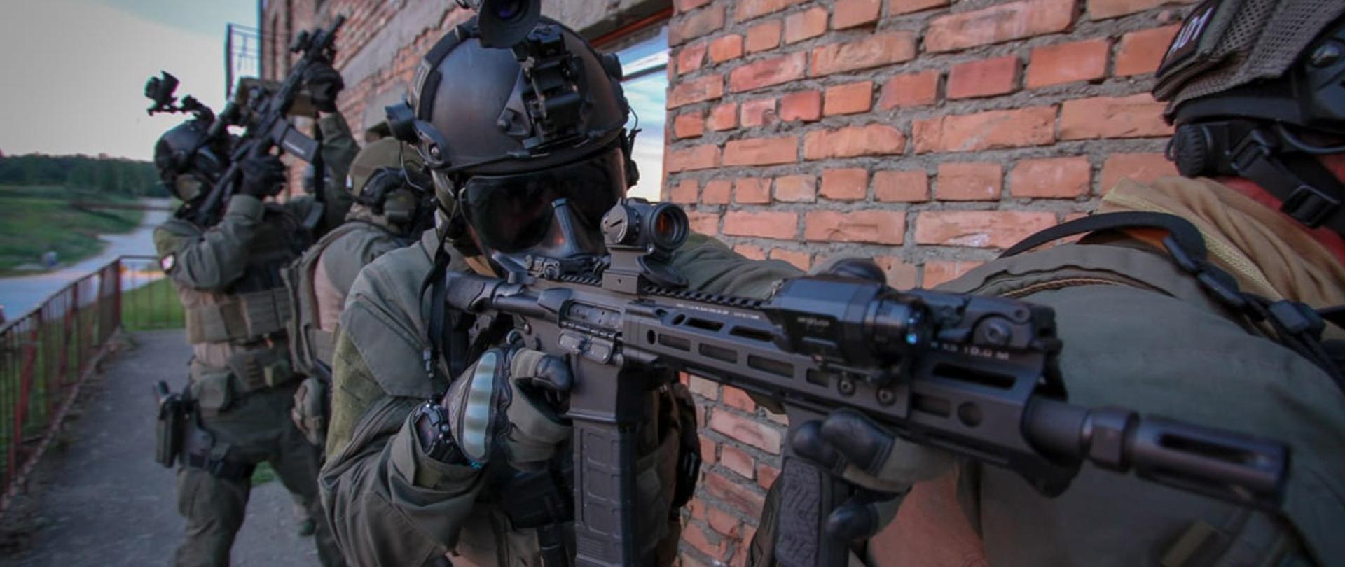 Uzbrojeni funkcjonariusze Służby Celno-Skarbowej podczas realizacji, w tle ceglasta ściana.