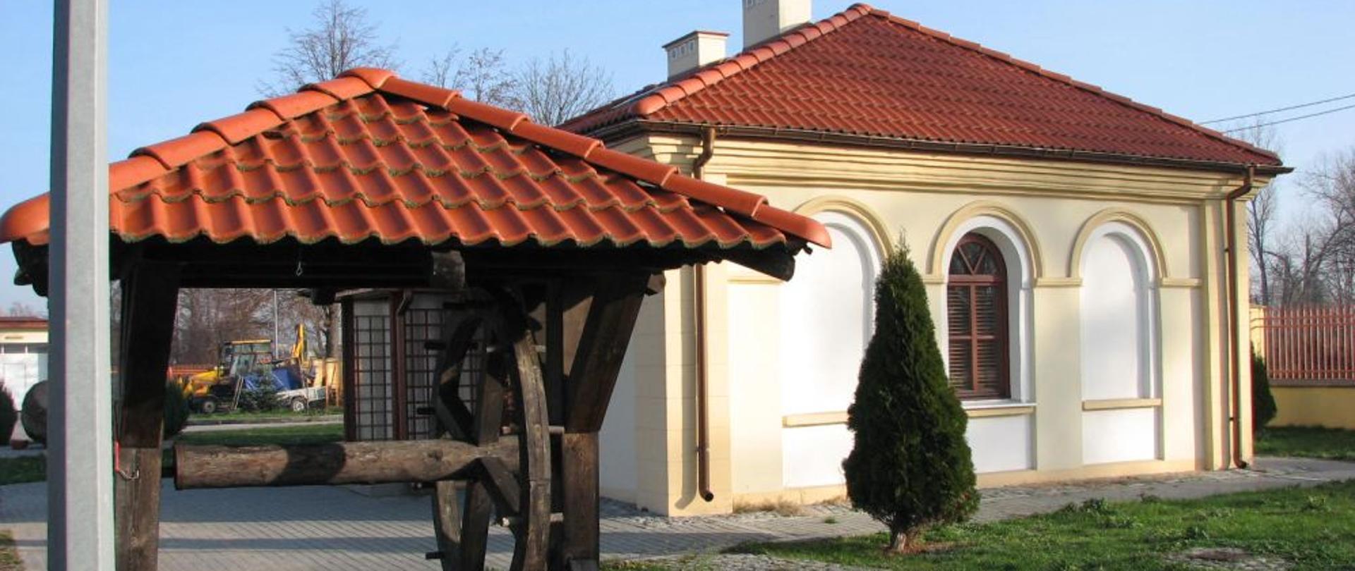 Dróżniczówka w Skarżysku-Kamiennej - na pierwszym planie stara studnia z drewnianym kołowrotkiem pod daszkiem z dachówek koloru ceglastego. Na drugim planie domek murowany koloru białego i kremowego z dachem spadzistym z ceglastej dachówki i z półkolistym oknem. Wokół zieleń.