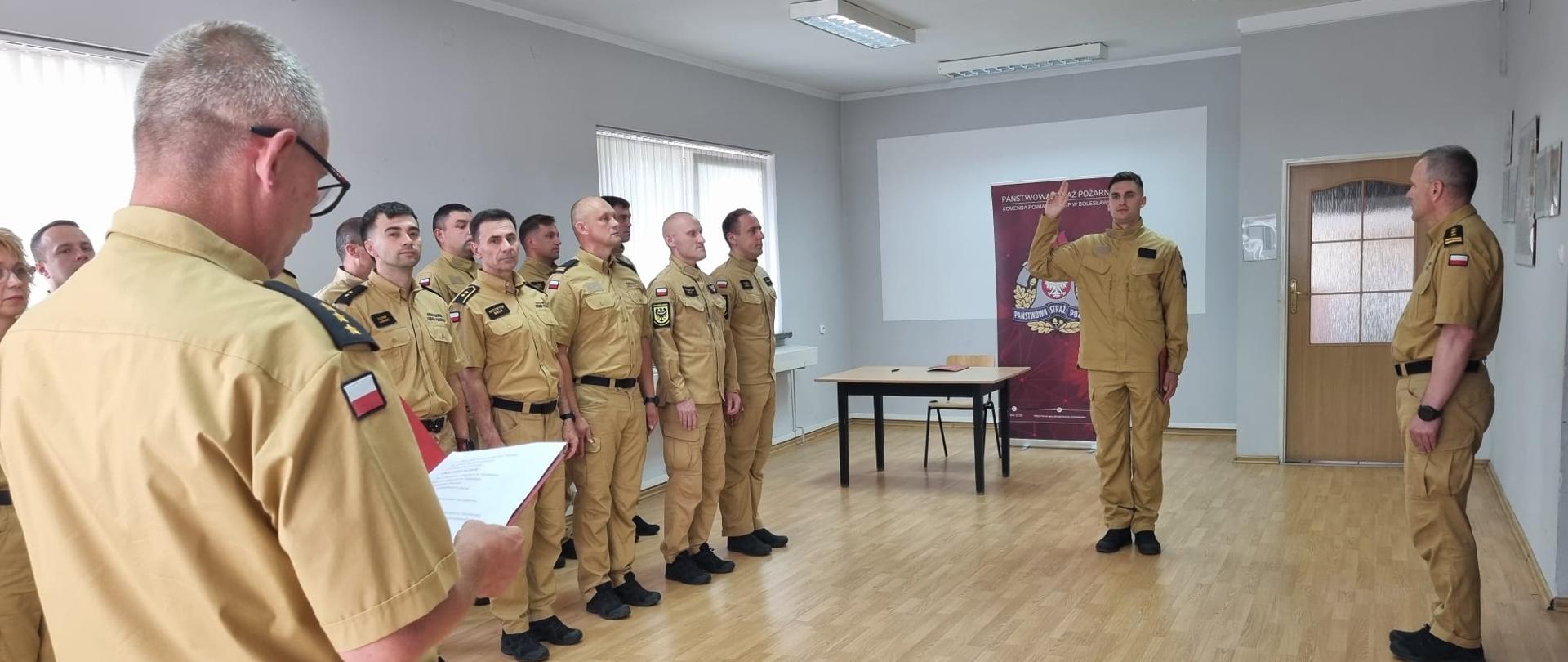 W sali szkoleniowej strażacy Państwowej Straży Pożarnej w umundurowaniu służbowym stoją w szeregu podczas uroczystości. Na pierwszym planie osoba odczytuje dokument, a jeden z funkcjonariuszy stojących przed grupą unosi prawą rękę do ślubowania. W tle znajduje się roll-up PSP oraz stół.