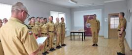 W sali szkoleniowej strażacy Państwowej Straży Pożarnej w umundurowaniu służbowym stoją w szeregu podczas uroczystości. Na pierwszym planie osoba odczytuje dokument, a jeden z funkcjonariuszy stojących przed grupą unosi prawą rękę do ślubowania. W tle znajduje się roll-up PSP oraz stół.