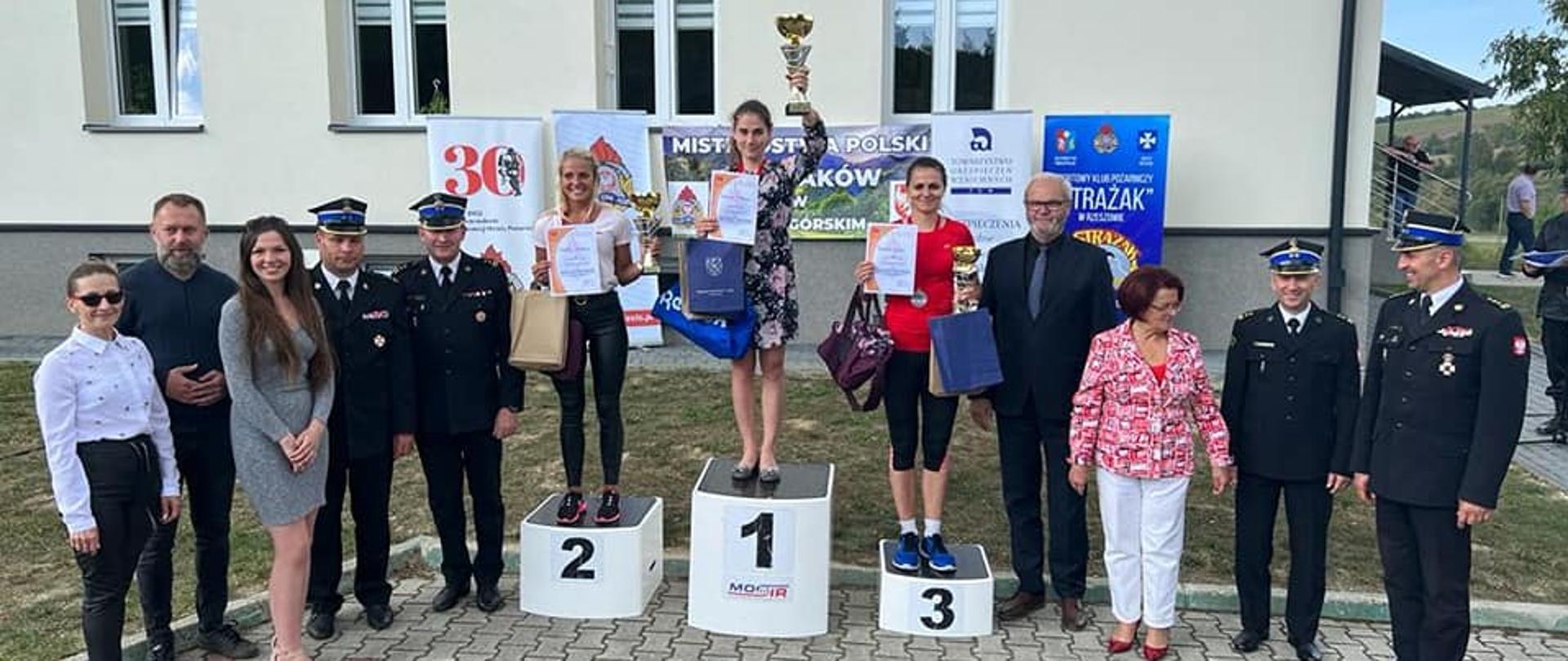 Zdjęcie przedstawia grupę osób stojących w łuku na utwardzonej nawierzchni przed budynkiem. Trzy kobiety w środku stoją na podium dla zwycięzców trzymając w rękach puchary i dyplomy.