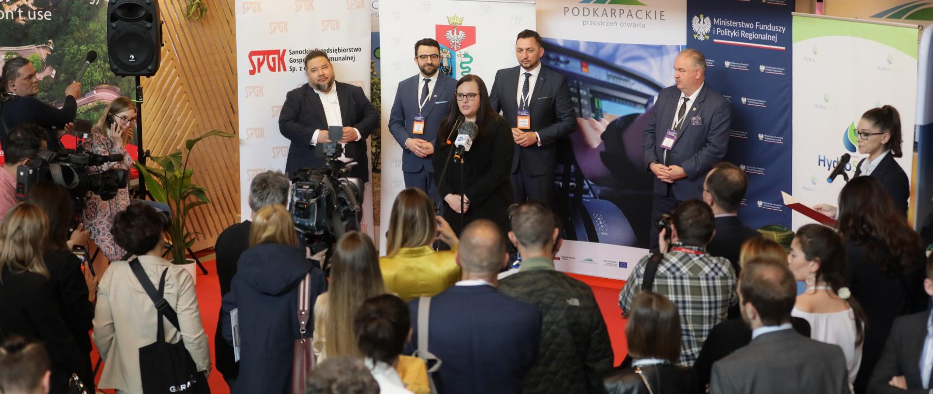 Pięć osób stoi na tle banerów podczas konferencji. Wiceminister Małgorzata Jarosińska-Jedynak przed mikrofonem. Wokół publiczność.