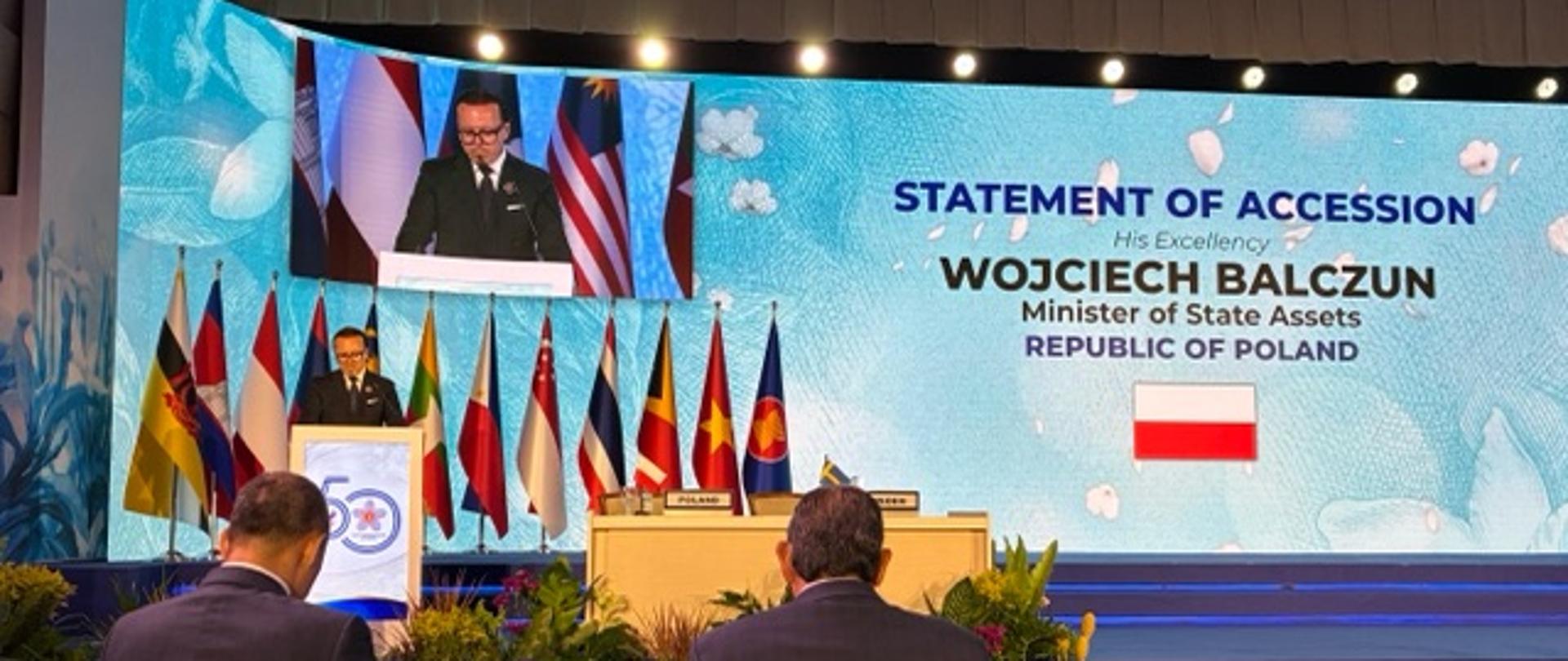 The photo shows a man in a suit. He is standing at the podium. Behind him are a dozen or so flags and a screen showing a live broadcast of his speech. Next to him is a wall with the words “Statement of Accession. His Excellency Wojciech Balczun. Minister of State for Assets. Republic of Poland.”