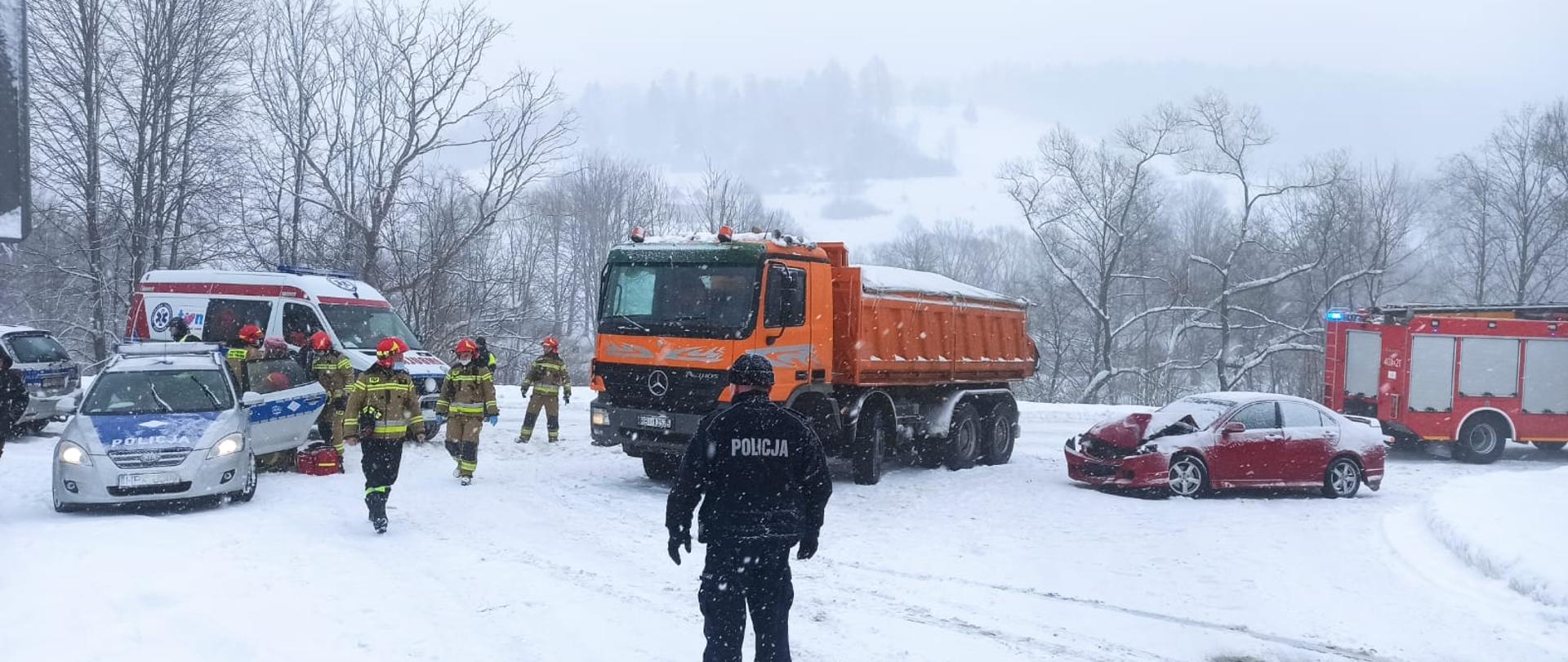 Na zdjęciu widać miejsce działań służb gdzie od lewej ustawiony jest samochód Policji za którym stoi karetka obok pojazdy biorące udział w zderzeniu, czyli ciężarowy samochód marki mercedes oraz samochód osobowy. Na końcu ustawiony jest średni samochód gaśniczy Państwowej Straży Pożarnej. Pojazdy te znajdują się na skrzyżowaniu dróg. Przy samochodzie Policji znajdują się strażacy którzy udzielają osobie poszkodowanej KPP oraz reszta która zabezpiecza miejsce zdarzenia. Przed ciężarówką stoi policjant. Aura jest zimowa a po drugiej stronie drogi znajdują się drzewa.