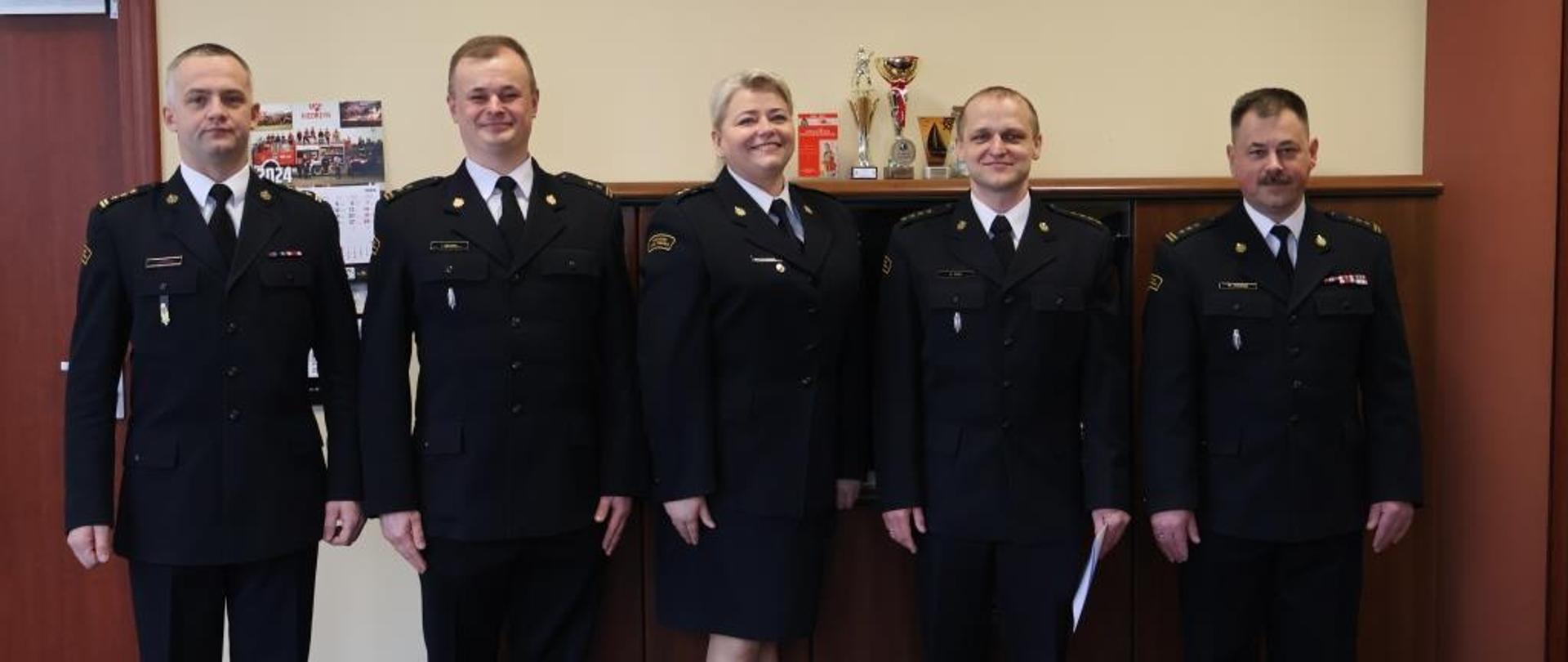 Zdjęcie wykonane w jednym z pomieszczeń komendy miejskiej Państwowej Straży Pożarnej w Radomiu. Na zdjęciu od lewej widać: pełniącego obowiązki komendanta miejskiego psp w Radomiu starszego brygadiera Łukasza Szymańskiego. Obok niego stoi dowódca jednostki ratowniczo – gaśniczej numer 2 w Radomiu. Następnie kolejno ustawieni są: zastępca naczelnika wydziału kadr, nowo mianowany na stanowisko zastępcy dowódcy jednostki numer 2 w Radomiu funkcjonariusz oraz zastępca komendanta miejskiego psp w Radomiu starszy brygadier Marcin Ziomek. Funkcjonariusze ubrani są w ubrania w mundury galowe w kolorze ciemnym granatowym.