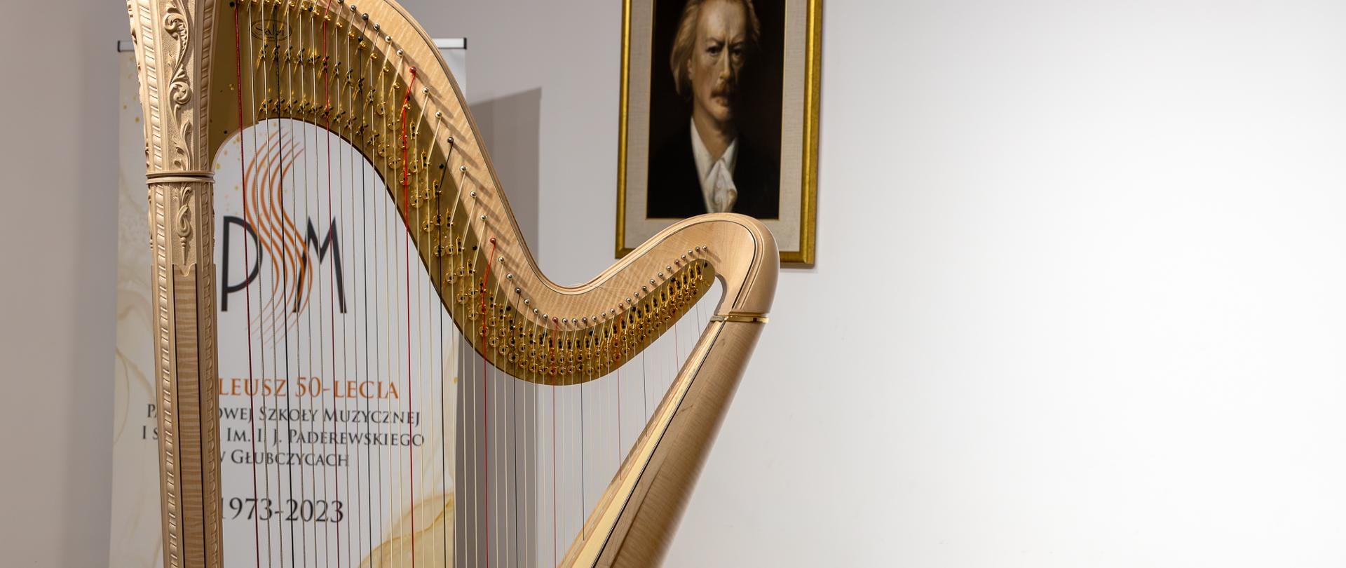 Harfa stojąca na scenie PSM w Głubczycach. W tle widoczny baner szkoły informujący o pięćdziesięcioleciu PSM oraz wiszący na ścianie portret patrona szkoły.