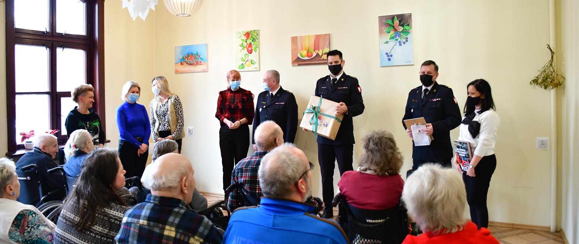 Pod ścianą w rzędzie stoją przedstawiciele Państwowej Straży Pożarnej oraz opiekunowie Domu Pomocy Społecznej. Przed nimi na krzesłach siedzą podopieczni DPS.