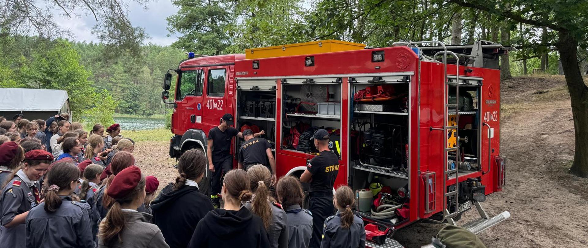 Zdjęcie przedstawia funkcjonariuszy KP PSP w Strzelcach Krajeńskich oraz grupę harcerzy ZHR, słuchających o zagrożeniach występujących podczas wypoczynku w sezonie letnim.