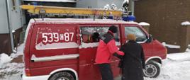 Strażak Ochotniczej Straży Pożarnej z Wielkiego Stwolna trzymając pod lewe ramię starszą Panią pomaga jej wsiąść do samochodu strażackiego typu bus.