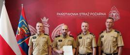 Na czerwony tle stoi 4 funkcjonariuszy Państwowej Straży Pożarnej w mundurach służbowych. Po lewej ich stronie stoją dwie flagi na stojakach, flaga Polski oraz miasta Szczecin. Od lewej mł. bryg. Marcin Grześkiewicz, mł. kpt. Emilian Skwarek, bryg. Jacek Cyburt, kpt. Tomasz Szmudziński.