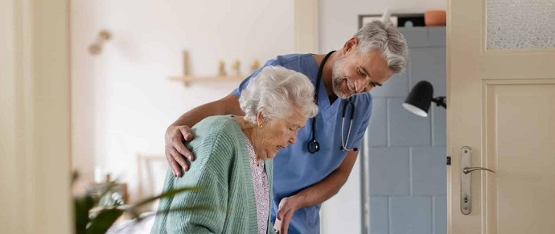 Grafika pokazuje seniora przemieszczającego się po domu opieki za pomocą balkonika; seniora wspomaga opiekun