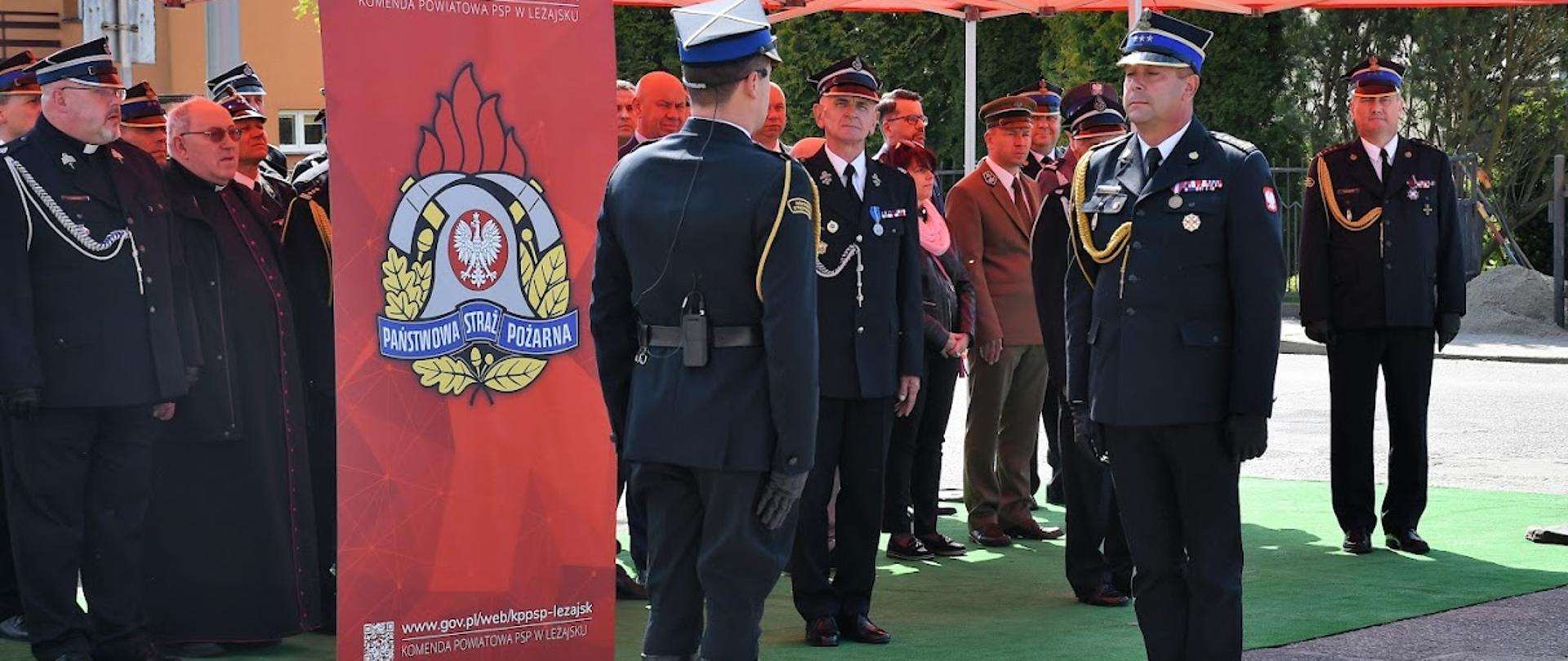 Zdjęcie zrobione na zewnątrz podczas uroczystości. Oficer Państwowej Straży Pożarnej w stopniu starszego brygadiera odbiera meldunek od dowódcy uroczystości. Za nimi, pod namiotem stoją zaproszeni goście. W tle widoczny jest rollup z napisem Państwowa Straż Pożarna Komenda Powiatowa Państwowej Straży Pożarnej w Leżajsku. Na rollup'ie umieszczono logotyp Państwowej Straży Pożarnej. 