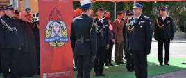 Zdjęcie zrobione na zewnątrz podczas uroczystości. Oficer Państwowej Straży Pożarnej w stopniu starszego brygadiera odbiera meldunek od dowódcy uroczystości. Za nimi, pod namiotem stoją zaproszeni goście. W tle widoczny jest rollup z napisem Państwowa Straż Pożarna Komenda Powiatowa Państwowej Straży Pożarnej w Leżajsku. Na rollup'ie umieszczono logotyp Państwowej Straży Pożarnej. 