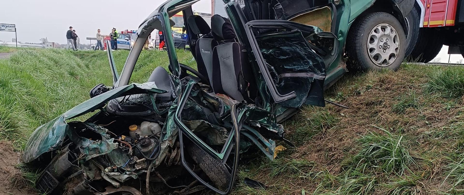 rozbity, zielony samochód osobowy po wypadku. Pojazd znajduje się na poboczu drogi, częściowo na trawie i ziemi, przechylony na bok. Przednia część samochodu jest doszczętnie zniszczona, widać zmiażdżony silnik i inne elementy mechaniczne. Drzwi po stronie kierowcy są otwarte i zniekształcone, podobnie jak inne części karoserii. Tylna klapa bagażnika jest podniesiona. Obok wraku stoi czerwony wóz strażacki, częściowo widoczny w prawym górnym rogu zdjęcia. W tle widać otwartą przestrzeń, prawdopodobnie pole lub łąkę, oraz kilka osób w oddali, w tym osoby w uniformach służb ratowniczych. Niebo jest pochmurne.