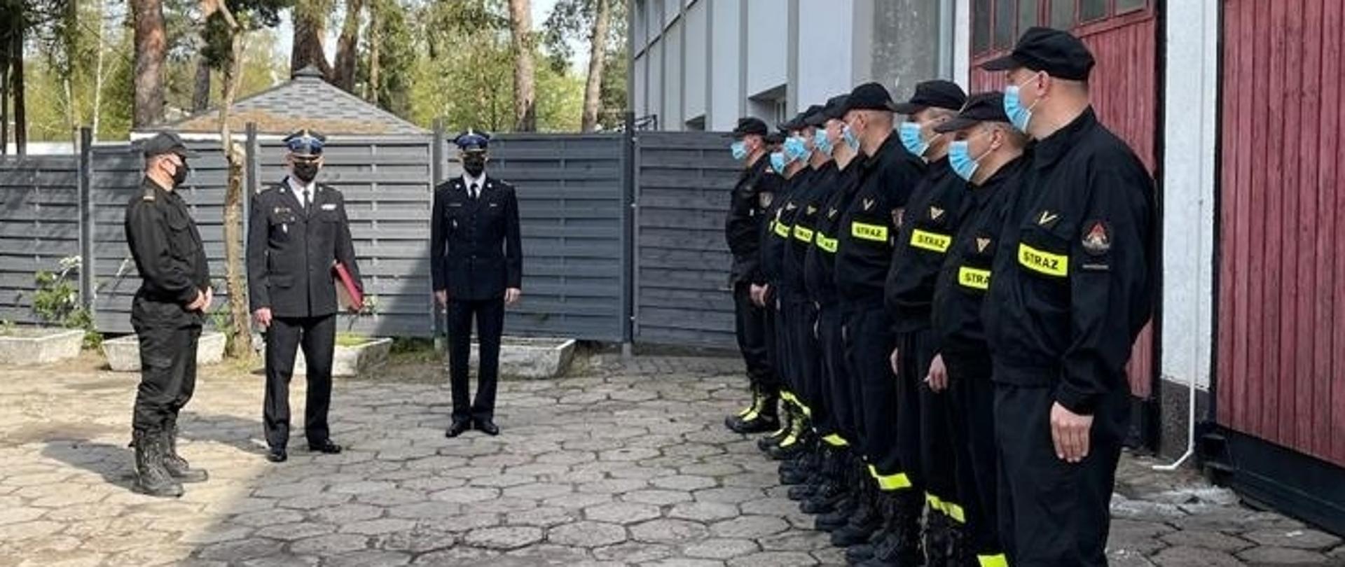 Komendant Miejski Państwowej Straży Pożarnej starszy brygadier Leszek Smuniewski ściska dłoń nowemu dowódcy Jednostki Ratowniczo Gaśniczej nr 14 młodszemu brygadierowi Ireneuszowi Żabiukowi. Obaj ubrani są w mundur wyjściowy. Na twarzach mają czarne maseczki. Komendant trzyma w lewym ręku czerwoną teczkę z aktem mianowania.