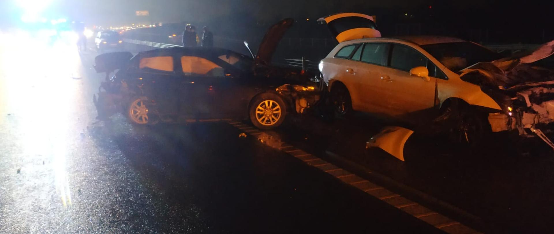 Zdjęcie zrobione w nocy na tle drogi ekspresowej. Na zdjęciu rozbite dwa samochody w wyniku wypadku drogowego. Po lewej stronie światła samochodów stojących w korku
