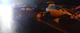 Zdjęcie zrobione w nocy na tle drogi ekspresowej. Na zdjęciu rozbite dwa samochody w wyniku wypadku drogowego. Po lewej stronie światła samochodów stojących w korku
