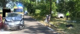 Widać uszkodzony samochód na drodze, za nim jest karetka pogotowia, po lewej rower na poboczu, w lesie samochód i policjant