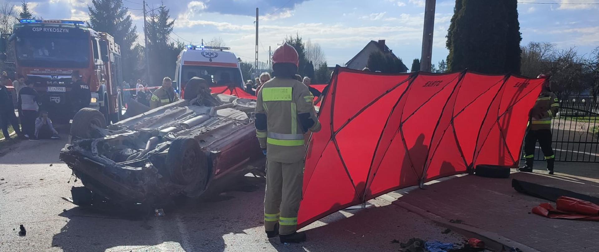Zdjęcie przedstawia ulicę Świętokrzyską. Po prawej stronie ustawiony został parawan osłaniający podtrzymywany przez dwóch strażaków. Na jezdni leży na dachu zniszczony samochód Opel Vectra, a w tle widać ambulans i wóz strażacki. 
