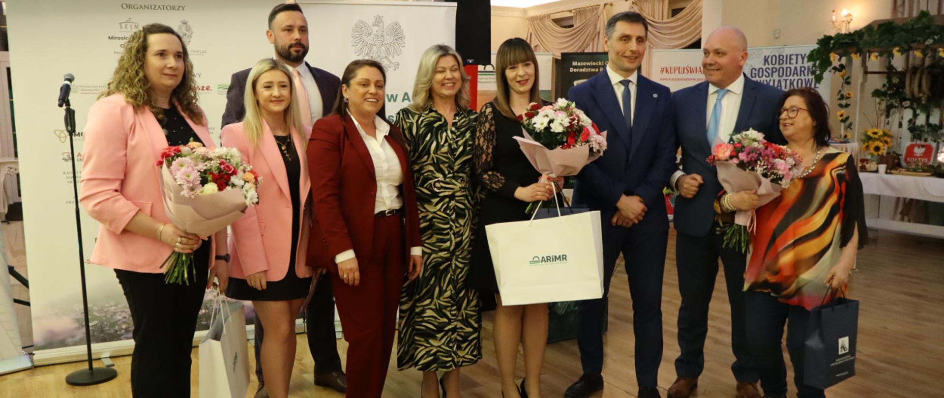 We wnętrzu eleganckiej Sali pozuje grupa sześciu kobiet i trzech mężczyzn. Wszyscy w galowych strojach, uśmiechnięci, w tle białe roll-upy