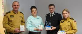 Cztery osoby stoją obok siebie w pomieszczeniu biurowym, pozując do zdjęcia. Dwie osoby po bokach oraz jedna po prawej stronie są ubrane w żółte mundury PSP, a jedna osoba w środku ma mundur OSP. Druga osoba od lewej ma na sobie jasnoniebieską bluzkę i czarne spodnie. Każda z osób trzyma w rękach pudełko z czujnikiem dymu. W tle widać jasne ściany, drzwi oraz roślinę doniczkową.