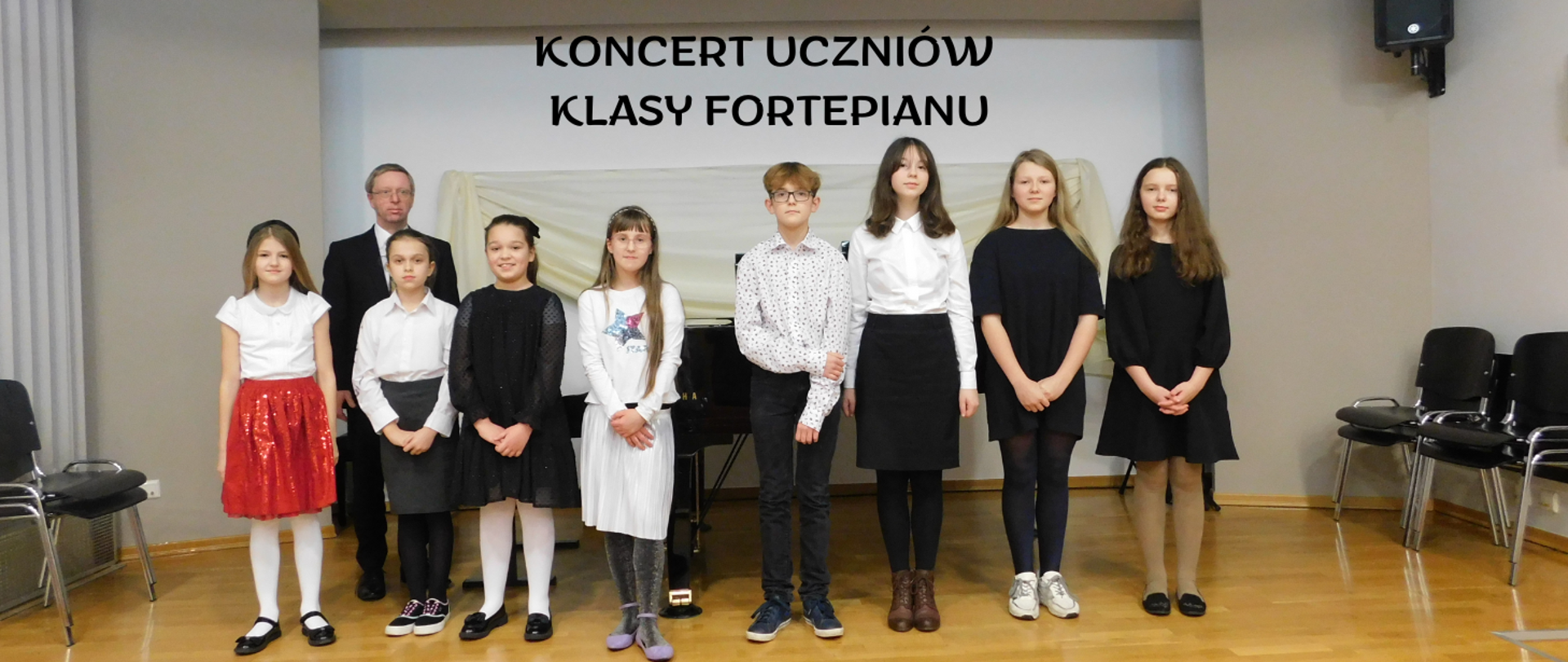 Na zdjęciu grupa uczniów, głównie dziewczynki i tylko jeden chłopiec. Wszyscy ubrani na galowo. Z tyłu za uczniami stoi mężczyzna w czarnej marynarce w okularach, obok niego czarny fortepian. U góry obrazka czarny napis "koncert uczniów klasy fortepianu".