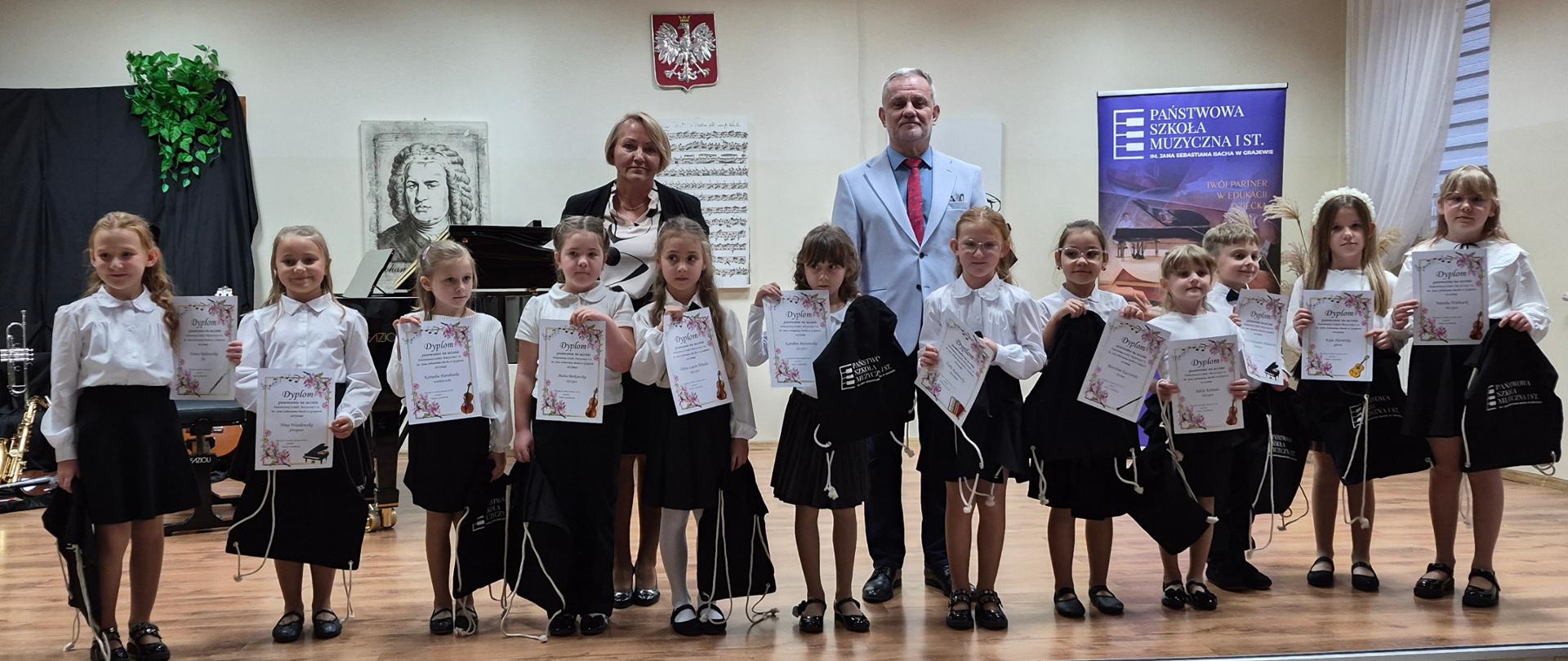 Uczniowie klas pierwszych cyklu sześcioletniego Państwowej Szkoły Muzycznej I stopnia im. Jana Sebastiana Bacha w Grajewie stoją na scenie z dyplomami, w towarzystwie nauczycieli. Dzieci ubrane są w stroje galowe(białe koszule i czarne spódnice/spodnie). W tle widoczne są fortepian, godło Polski oraz baner szkoły