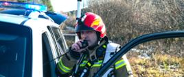 Na zdjęciu widać Dowódcę JRG w ubraniu specjalnym przy samochodzie operacyjnym z włączonymi światłami błyskowymi. Dowódca rozmawia przez radiotelefon.