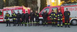 zbiórka w szeregu strażaków na tle dwóch samochodów ratowniczo-gaśniczych