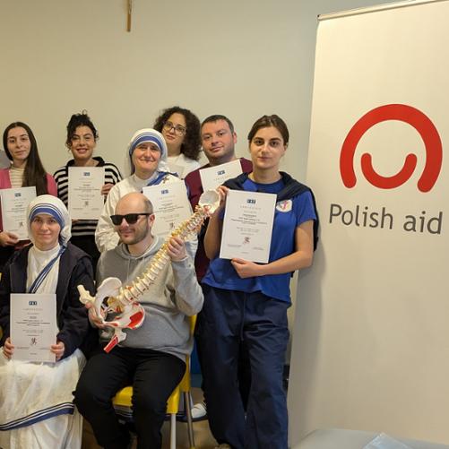 Grupa kobiet i mężczyzn pozuje z certyfikatami, z przodu siedzą dwie osoby, z których jednak ma model szkieletu dłoni a druga szkieletu kręgosłupa i miednicy