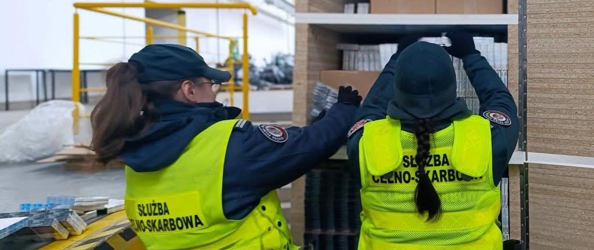 Dwie osoby w kamizelkach z napisem „Służba Celno‑Skarbowa” stoją przy metalowym regale i sprawdzają ułożone na półkach opakowania; w tle widać elementy hali magazynowej oraz żółtą barierkę.