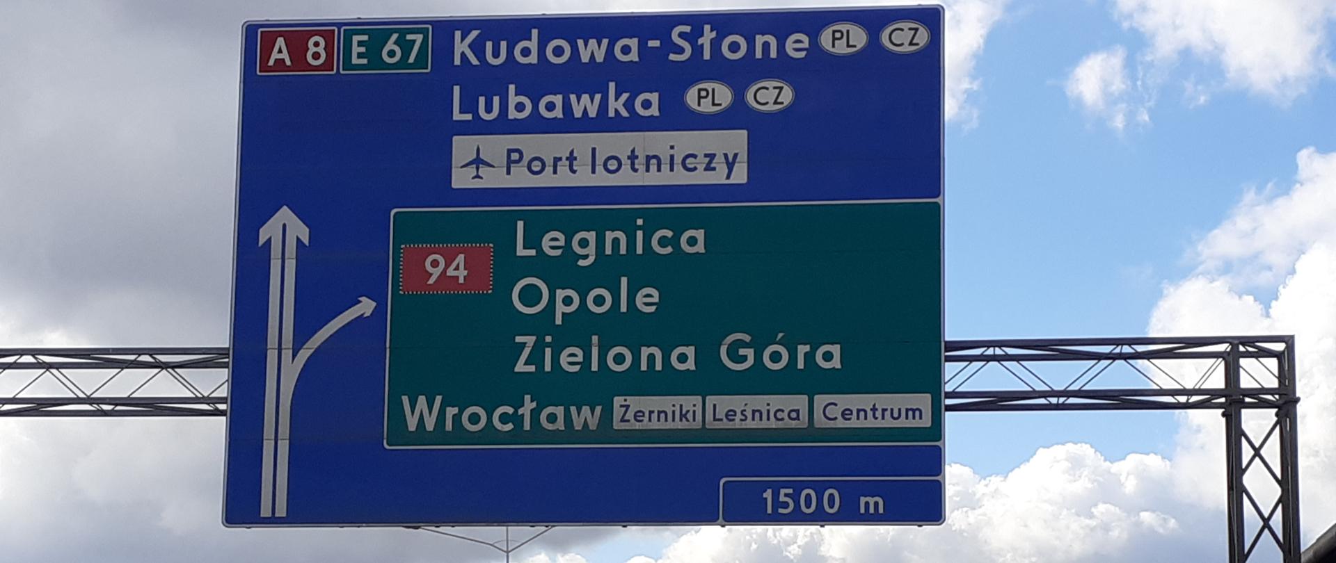 na zdjęciu widoczna autostrada nad którą widnieje duża tablica drogowskazowa. Po autostradzie poruszają się samochody 