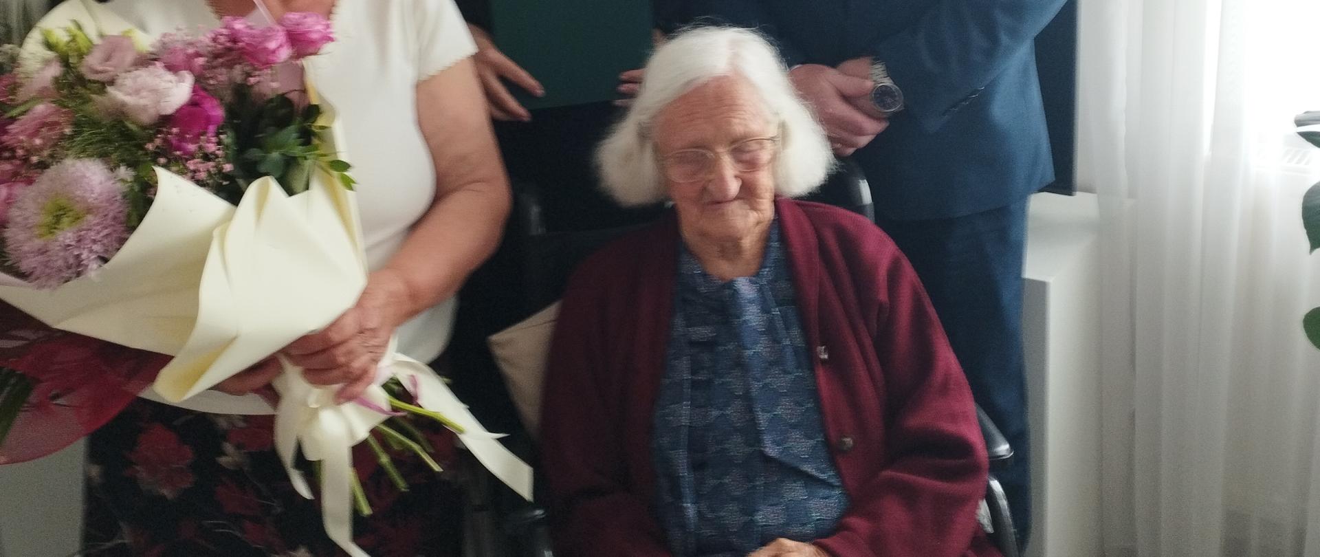 W pokoju na wózku siedzi starsza kobieta z długimi białymi włosami - Jubilatka Pani Eufemia Gadomska. Obok po lewej stronie stoi młodsza kobieta z siwymi upiętymi włosami w białej bluzce - córka Jubilatki, która trzyma w rękach bukiet pięknych kwiatów. Za Jubilatką stoją mężczyzna i kobieta, która trzyma w rękach list gratulacyjny w zielonej okładce ze złotym logo KRUS. Jubilatka uśmiecha się do zdjęcia przejęta życzeniami i gratulacjami.