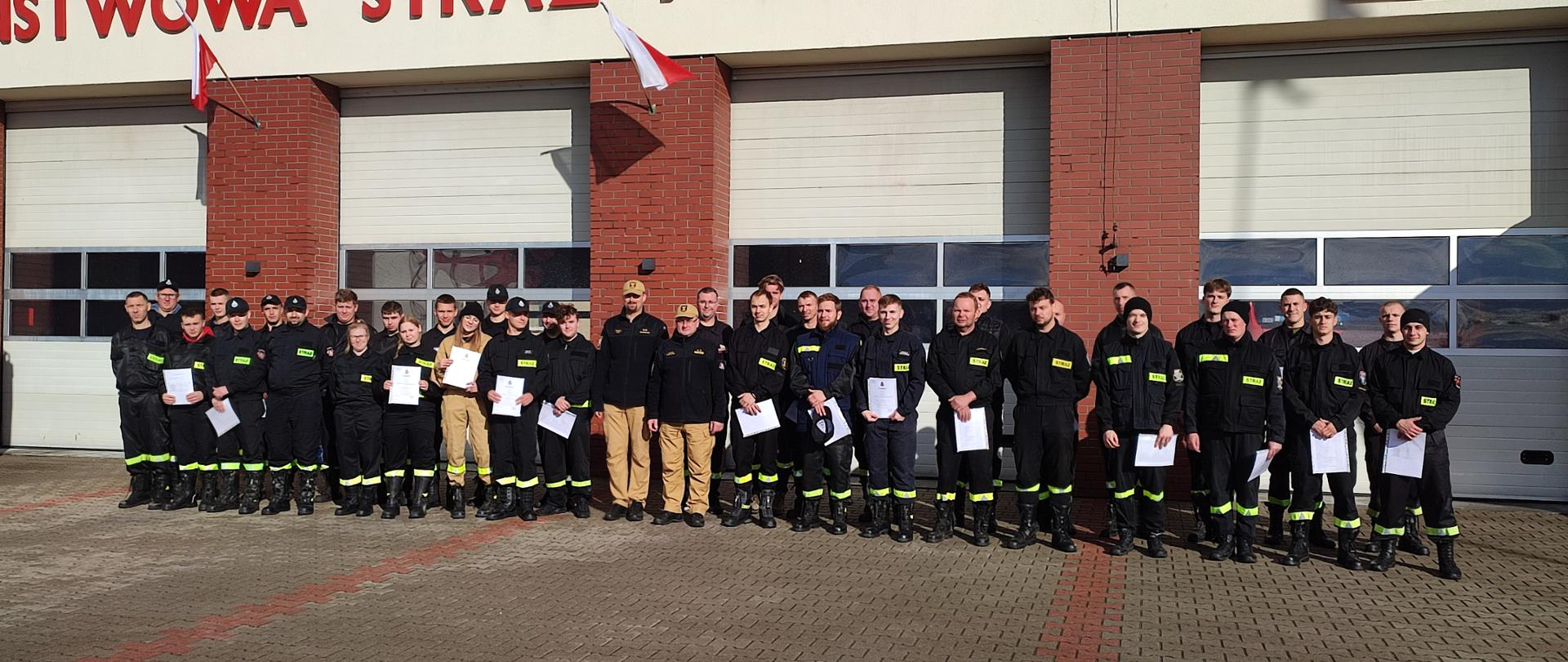 Absolwenci szkolenia podstawowego strażaków ratowników OSP wraz z Komendantem Powiatowym PSP w Środzie Wlkp. i Dowódcą JRG na placu przed budynkiem komendy w słoneczny dzień.