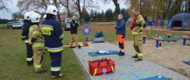 Warszaty szkoleniowe. Zdjęcie przedstawia fragment stadionu w Sarnowie, na którym przygotowane jest stanowisko do udzielania kwalifikowanej pierwszej pomocy. Na chodniku z kostki brukowej znajdują sięfantomy, deska ortopedyczna oraz trorba ze sprzętem medycznym. Wokół stanowiska znajdują sie instruktorzy oraz grupa strażaków biorących udział w warsztatach. Jest tam też zastępca komendanta powiatowego PSP. Część strażaków ma na sobie ubrania specjalne koloru piaskowego, część - ciemnego, a także hełmy w kolorze białym. 