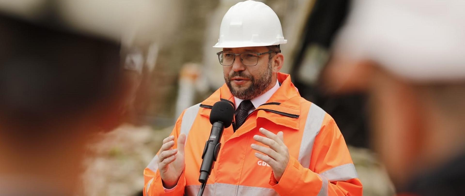 Minister funduszy i polityki regionalnej Grzegorz Puda stoi przy mikrofonie i przemawia, minister ubrany jest w kask i kamizelkę ochronną