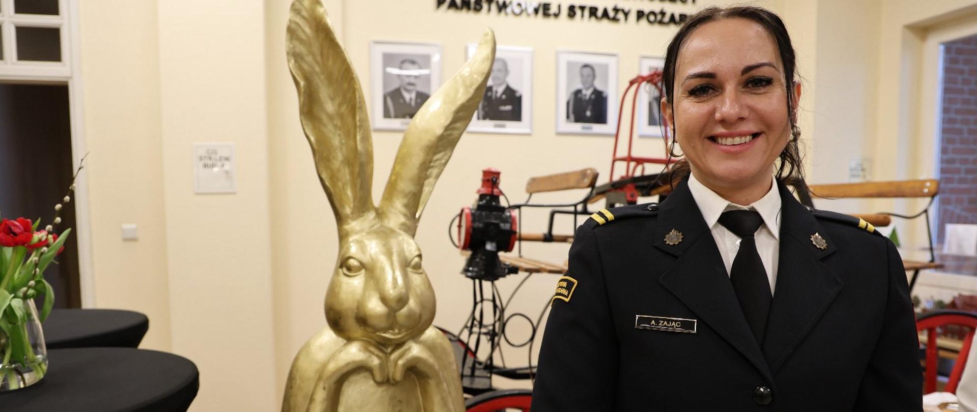Wnętrze jasnej sali. Na pierwszym planie stoi uśmiechnięta kobieta w ciemnym mundurze strażackim z białą koszulą i czarnym krawatem. Ma związane włosy i przypiętą plakietkę z nazwiskiem. Obok niej, po lewej stronie, znajduje się duża złota rzeźba królika z długimi uszami ustawiona na stole. Na stole widać także stos białych talerzy i dekoracje. W tle na ścianie wiszą czarno-białe portrety oraz napis w języku polskim. Za kobietą znajduje się zabytkowy sprzęt strażacki.