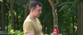 Funkcjonariusz trzyma gaśnicę i pokazuje jej obsługę, wskazując elementy urządzenia; w tle znajdują się drzewa i przygotowane miejsce do ćwiczeń.
Przekaż swoją opinię na temat programu.