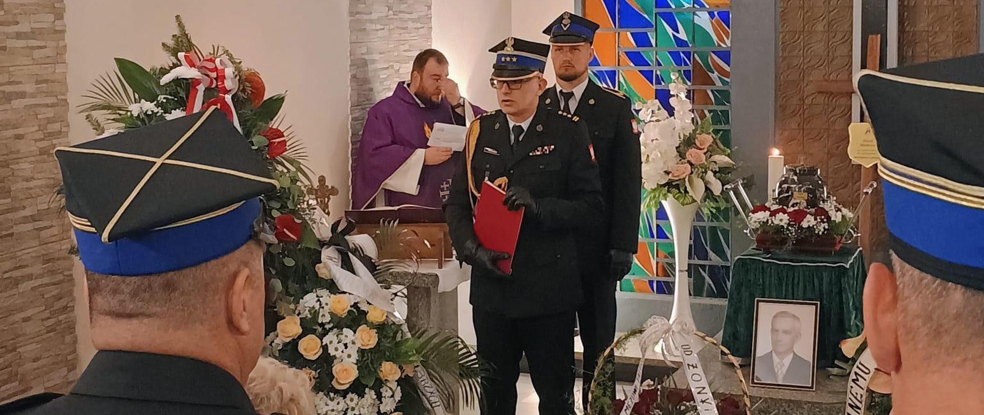 Zastępca komendanta głównego PSP przemawia do osób zgromadzonych na ceremonii pogrzbowej. Widoczna na katafalku urna z prochami zmarłego oraz jego zdjęcie.