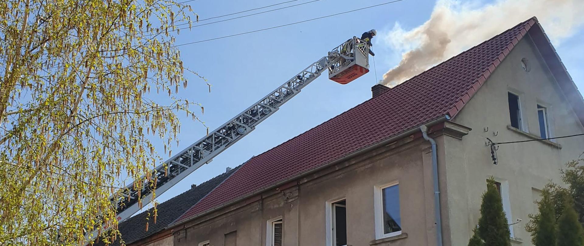 Pożar sadzy w kominie w miejscowości Pogorzela - zdjęcie przedstawia strażaka pracującego w koszu drabiny mechanicznej, która znajduje się nad dachem przy kominie budynku w którym doszło do pożaru. Dach jest w kolorze czerwonym.