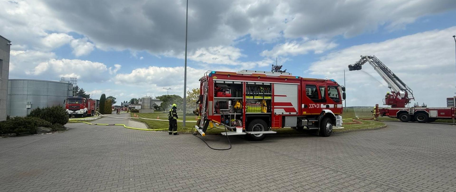 Na terenie zakładu, przed halami znajdują się rozstawione pojazdy pożarnicze pomiędzy którymi poprowadzono linie gaśnicze z węży strażackich. W tle znajduje się również rozstawiony podnośnik strażacki. Strażacy ćwiczą.