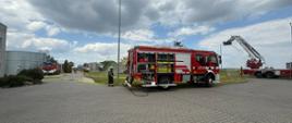 Na terenie zakładu, przed halami znajdują się rozstawione pojazdy pożarnicze pomiędzy którymi poprowadzono linie gaśnicze z węży strażackich. W tle znajduje się również rozstawiony podnośnik strażacki. Strażacy ćwiczą.